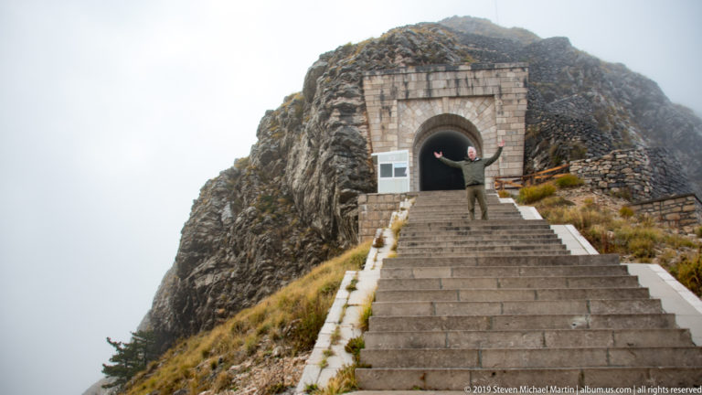 Broad stone steps climbing to an arched tunnel entrance set in a rocky knoll, with a visitor near the portal.