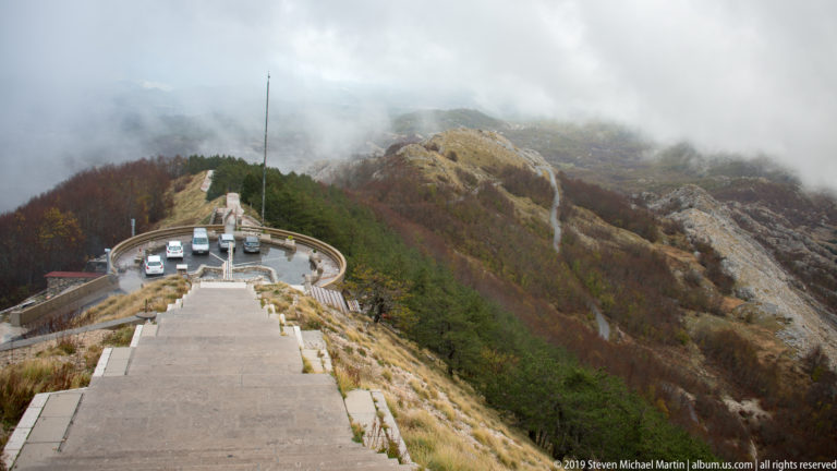 Long stone staircase descending a mountain ridge toward a small parking loop, vehicles, autumn forest, and foggy rocky slopes.