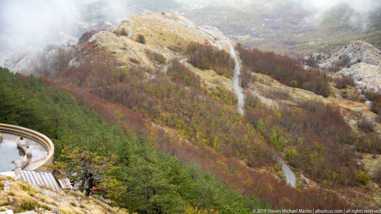 Winding pale road cutting through red, yellow, and green autumn woodland on a rocky Lovćen mountainside.