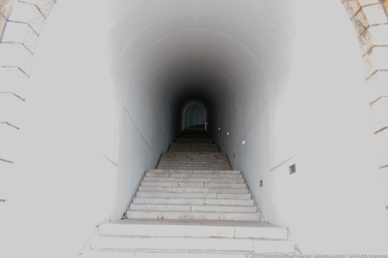 Long white tunnel with stone stairs and evenly spaced wall lights converging toward a distant arched opening.