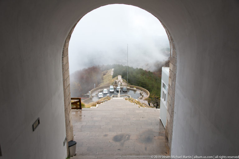 View through an arched tunnel exit down broad stone steps to a small parking area and foggy mountain slopes.