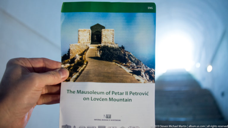 Hand holding an English brochure titled “The Mausoleum of Petar II Petrović on Lovćen Mountain” inside the stair tunnel.