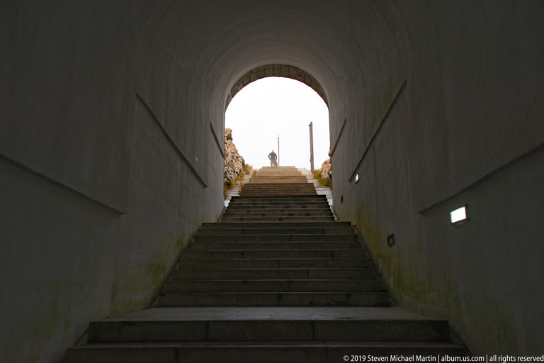 Stone steps ascending through a dim tunnel toward a bright arched exit, with small rectangular wall lights along both sides.