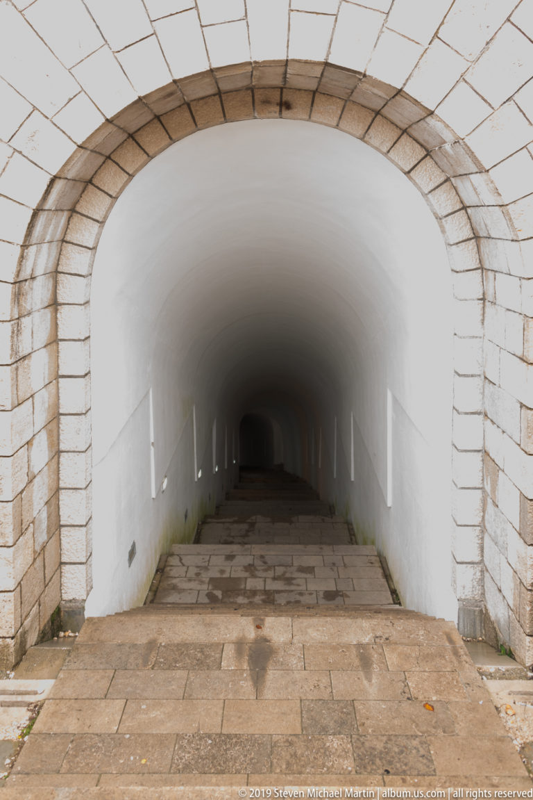 Steep stone stairway descending through a long white barrel-vaulted tunnel with small wall lights fading into darkness.