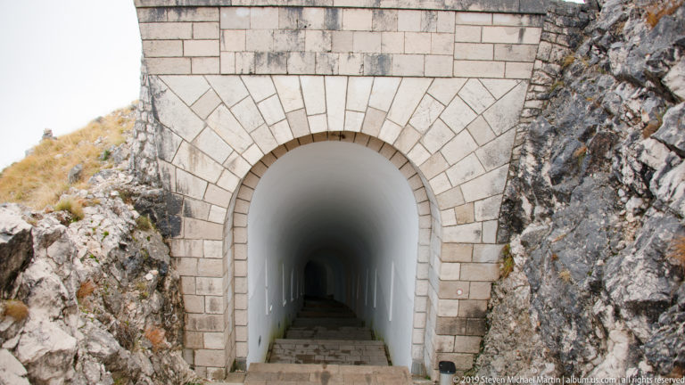 Rounded stone portal set into rock, opening onto a long white-walled stair tunnel descending into darkness.