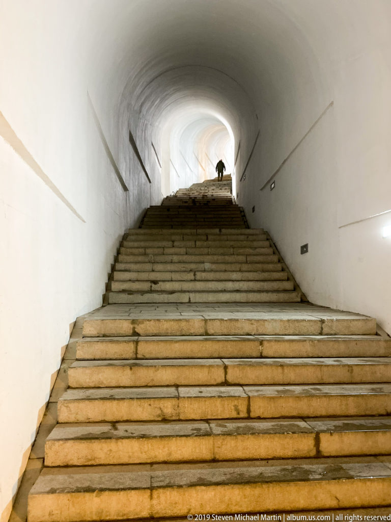 Wide stone steps rising through a white vaulted tunnel toward a silhouetted visitor at the bright upper landing.