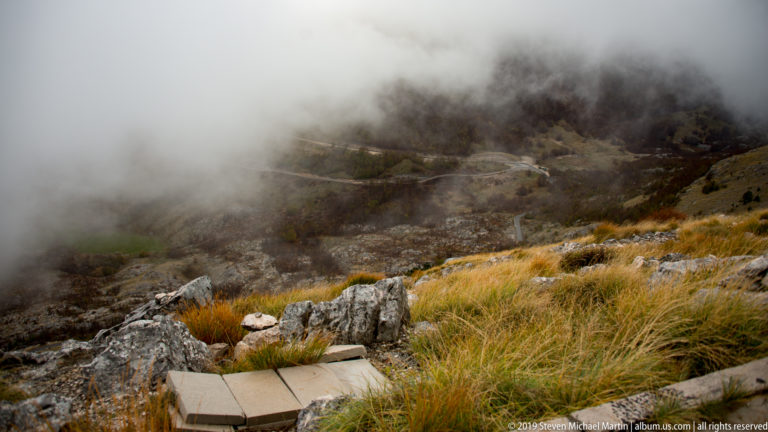 Fog moving over a rocky limestone slope with golden grass, scrub, and a faint road or path in the distance.
