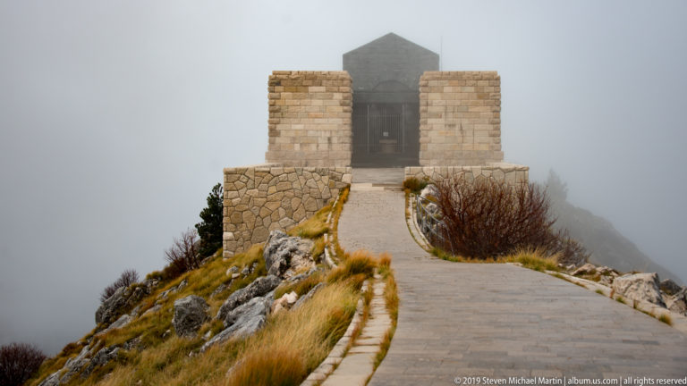 Fog-shrouded Njegoš Mausoleum at the end of a narrow paved ridge path bordered by ochre grass and rocks.