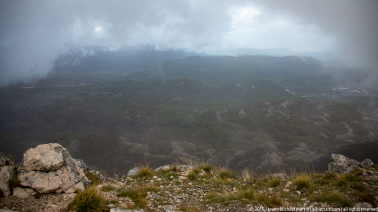 Rocky alpine foreground overlooking dark mountain ridges partly hidden by dense gray mist.