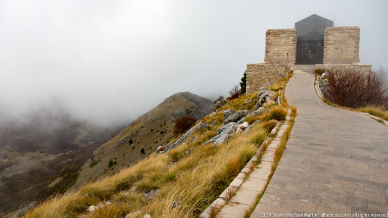 Stone path leading along a misty rocky ridge to the block-like Njegoš Mausoleum on the summit.