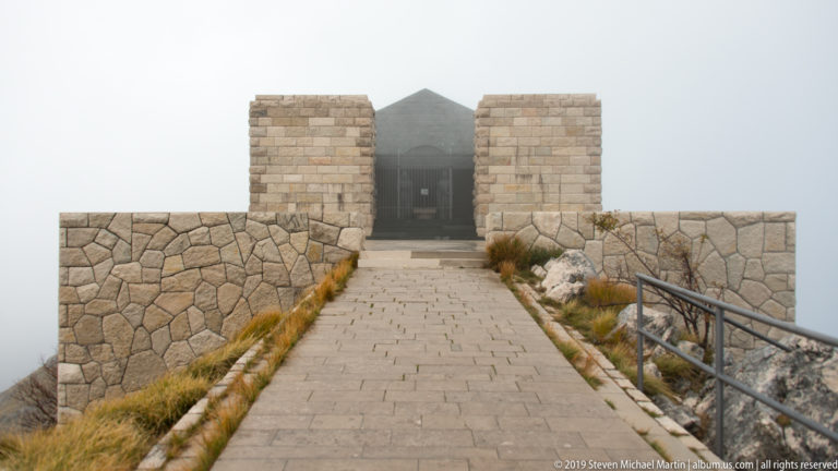 Paved ridge walkway leading through low stone walls toward the twin entrance pylons of the fog-shrouded Njegoš Mausoleum.