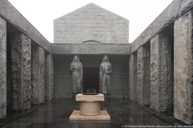 Symmetrical stone forecourt with a circular central element and two monumental female statues flanking the entrance beyond.