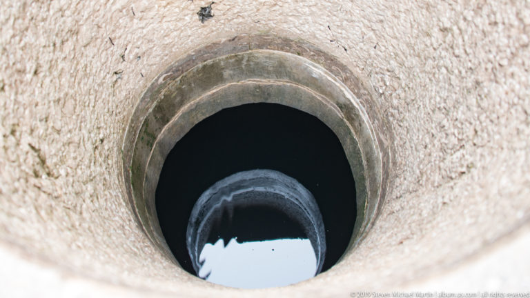 Top-down view into a circular stone-lined opening with dark interior and a bright reflection on water at the bottom.