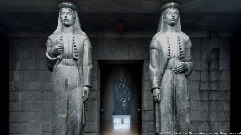 Two monumental gray stone women flank a dark doorway, with the seated Njegoš statue visible in the chamber beyond.