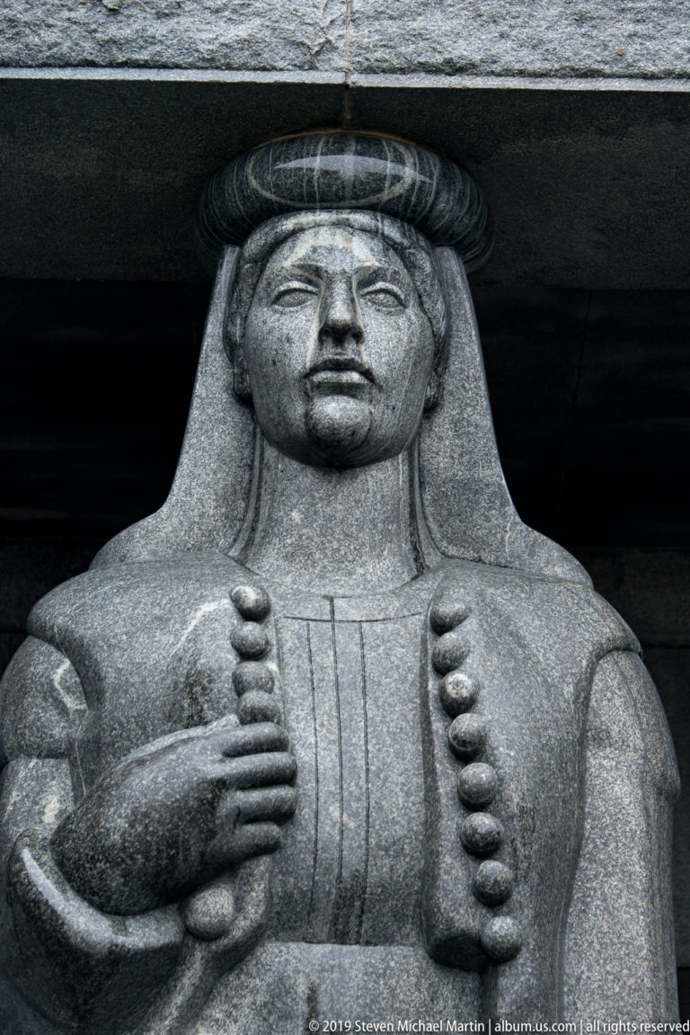 Close view of a monumental gray stone female figure with long stylized hair and a patterned garment.
