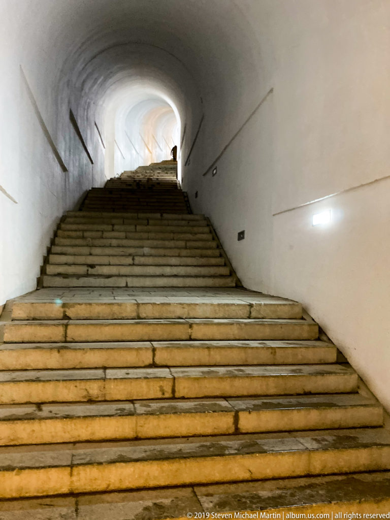 Broad stone staircase rising through a white vaulted tunnel toward a bright upper landing with one small silhouetted figure.