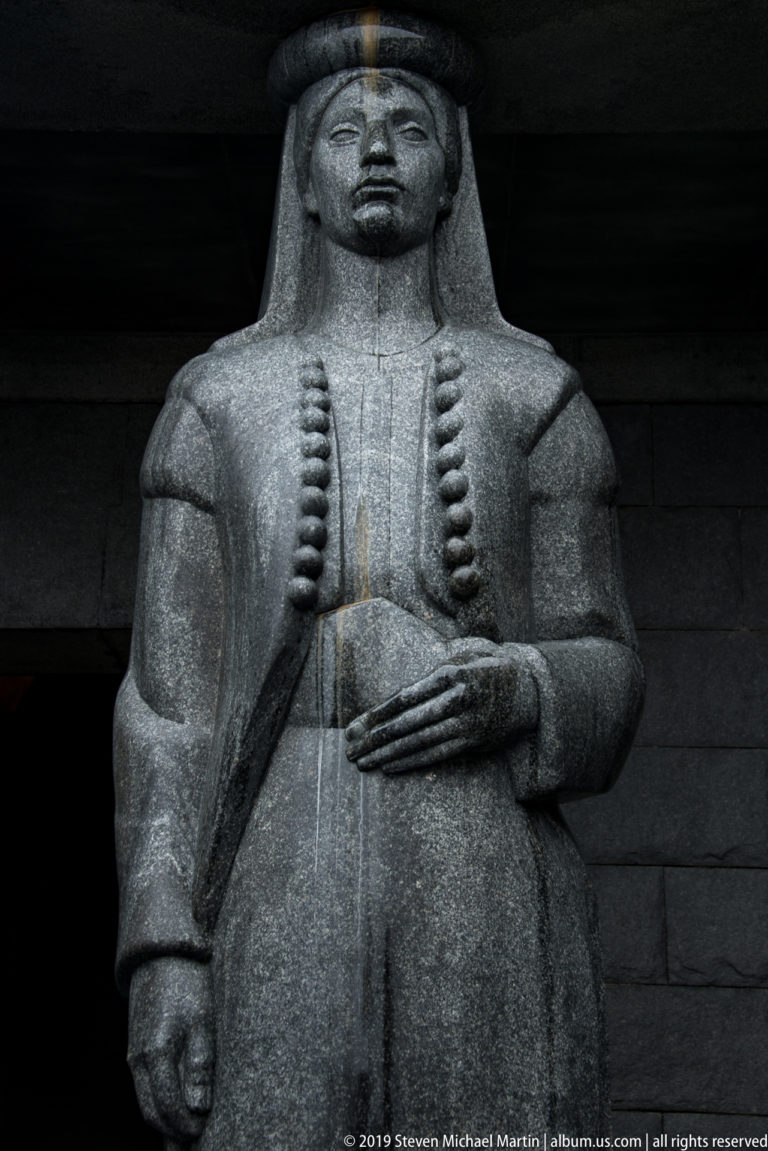 Monumental gray stone woman in traditional dress, shown frontally with long braided ornaments and one hand across her abdomen.