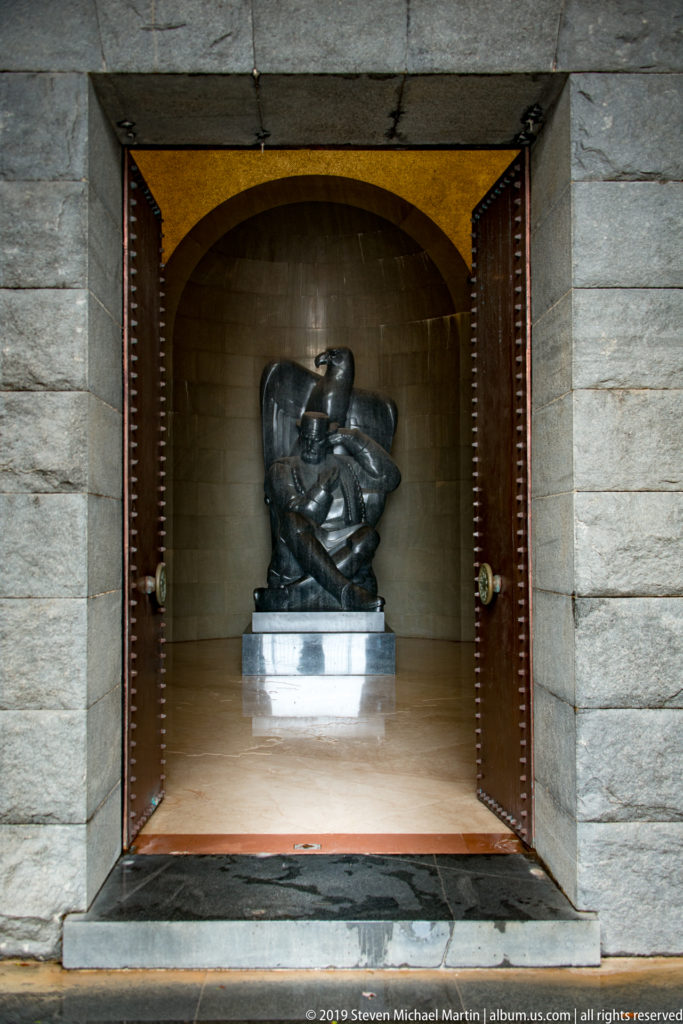 Dark sculptural group on a pale pedestal inside an arched stone chamber, seen through open studded doors.