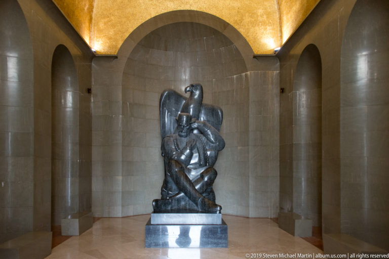 Dark seated statue of Petar II Petrović-Njegoš centered in a pale stone chamber beneath a glowing gold-toned ceiling.