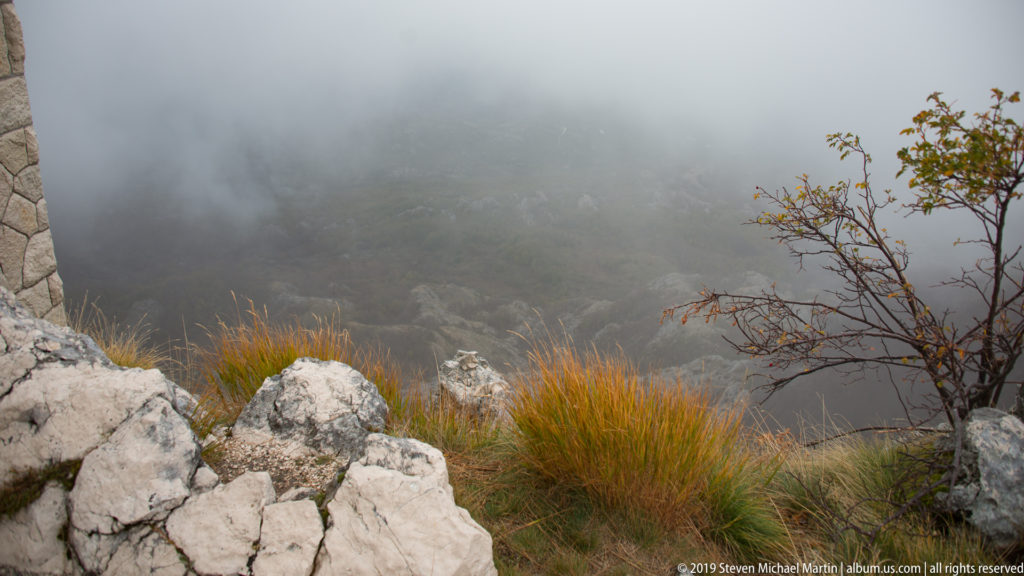 White limestone rocks and ochre alpine grass on a steep foggy slope, with a small autumn-leaved shrub at right.