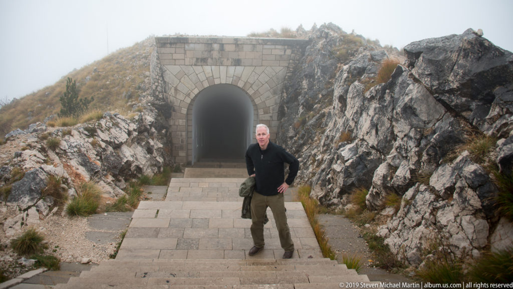 Visitor standing on stone paving before an arched masonry tunnel entrance set between rocky outcrops.