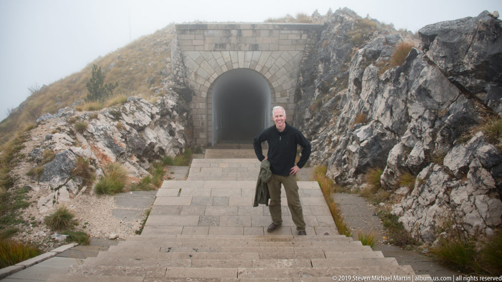 Wide view of stone steps climbing between limestone walls to an arched tunnel entrance, with a visitor standing below.