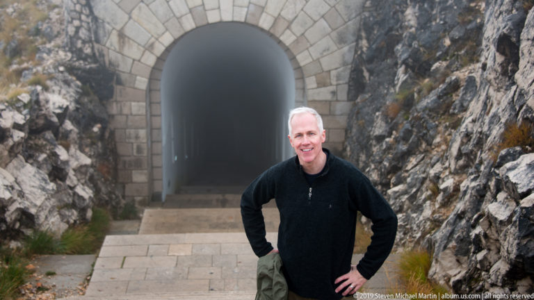 Man standing before a large arched stone tunnel entrance cut between steep rocky walls on the Lovćen summit approach.