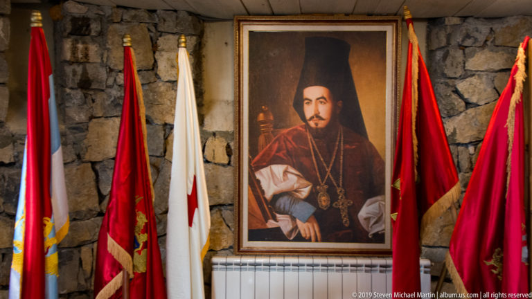 Framed portrait of a bearded Orthodox cleric in black headwear and red vestments, flanked by ceremonial flags.