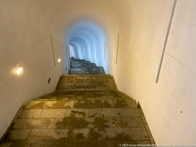 Worn stone stairs descending through a white vaulted tunnel with wall lights and a cooler blue-lit section below.