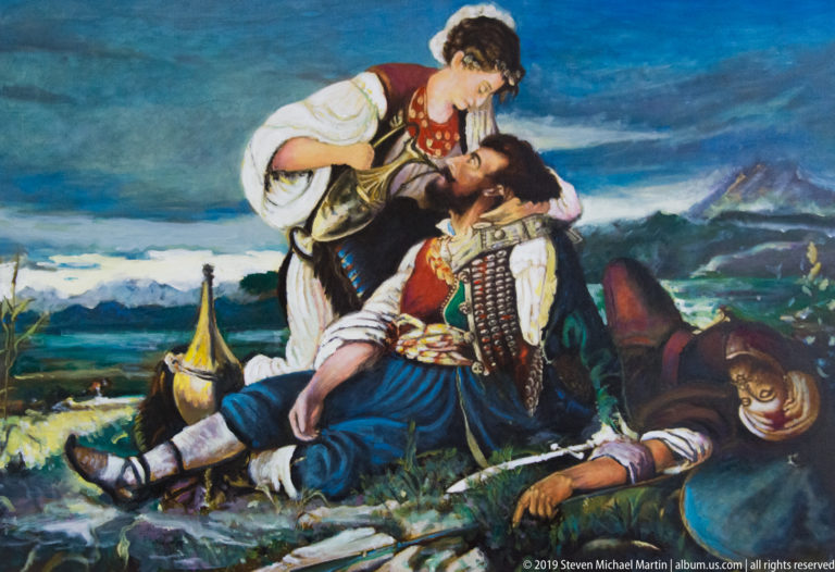 Woman in traditional dress bends over a wounded man in a dramatic mountain scene with another fallen figure nearby.