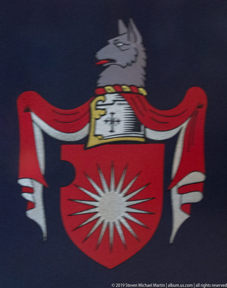 Red heraldic shield with white starburst, helmet, gray wolf crest, and red-and-white mantling on dark fabric.
