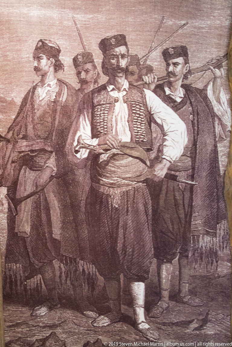 Sepia historical print of armed men in traditional dress, centered on a standing figure with sash and patterned vest.