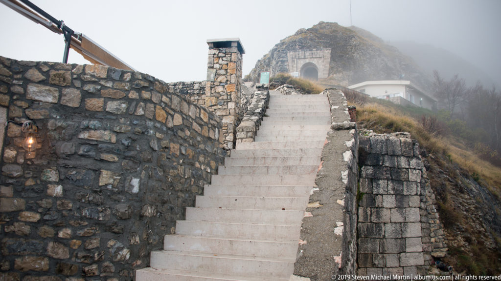 Steep pale staircase between stone walls climbing toward a misty arched passage near the Njegoš Mausoleum.