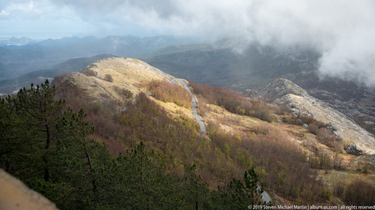 Sunlit rocky ridge with tawny grass and rust-colored woodland, framed by dark pines beneath low cloud.