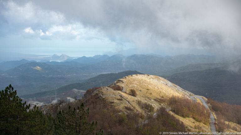 Rounded sunlit mountain ridge with autumn woodland before darker distant ranges beneath heavy storm clouds.
