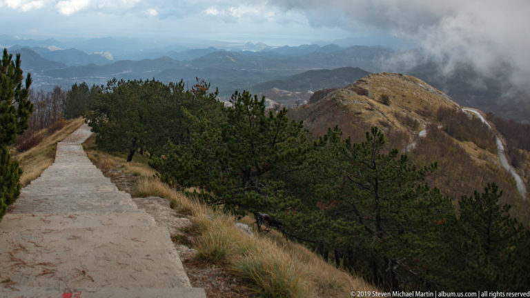 Paved ridge path beside low pines and dry grass, overlooking layered mountains and valleys under drifting clouds.