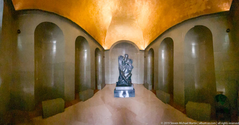 Long pale stone chamber with repeated arched recesses and a glowing gold-toned ceiling leading to the distant Njegoš statue.