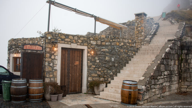 Stone visitor building with wooden doors and barrels beside a steep pale stairway climbing toward the misty summit.