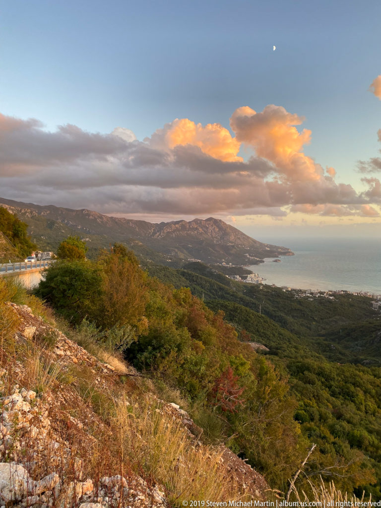 High mountain roadside view across wooded slopes to the Adriatic coast beneath warm evening clouds.
