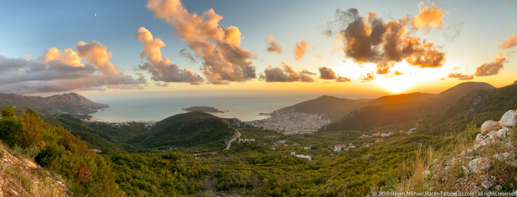 Sunset panorama from high mountains toward the Adriatic, with forested ridges, a coastal settlement, sea, and winding road.
