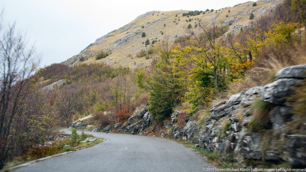 Paved mountain road curving between exposed limestone and autumn trees on the Lovćen approach.