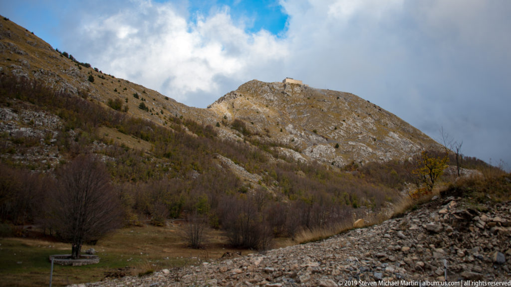 Wide view of a rocky Lovćen summit with the small Njegoš Mausoleum on the crest above leafless lower slopes.