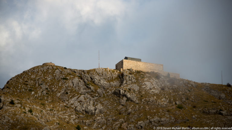Block-like Njegoš Mausoleum crowning a rocky Lovćen summit beneath low cloud, with a slender mast at right.