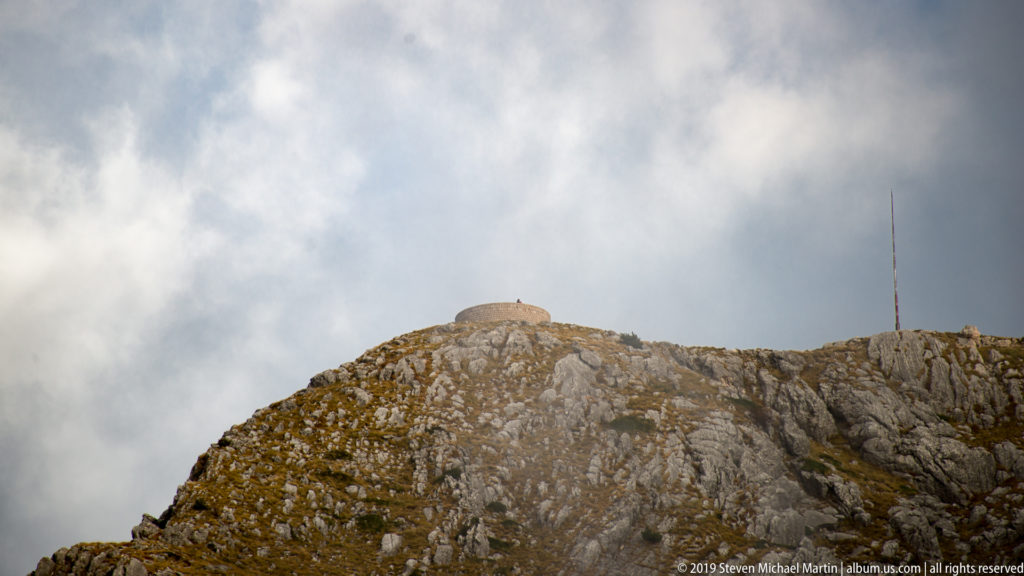 Jagged limestone summit ridge with sparse grass and a slender antenna rising into drifting cloud.