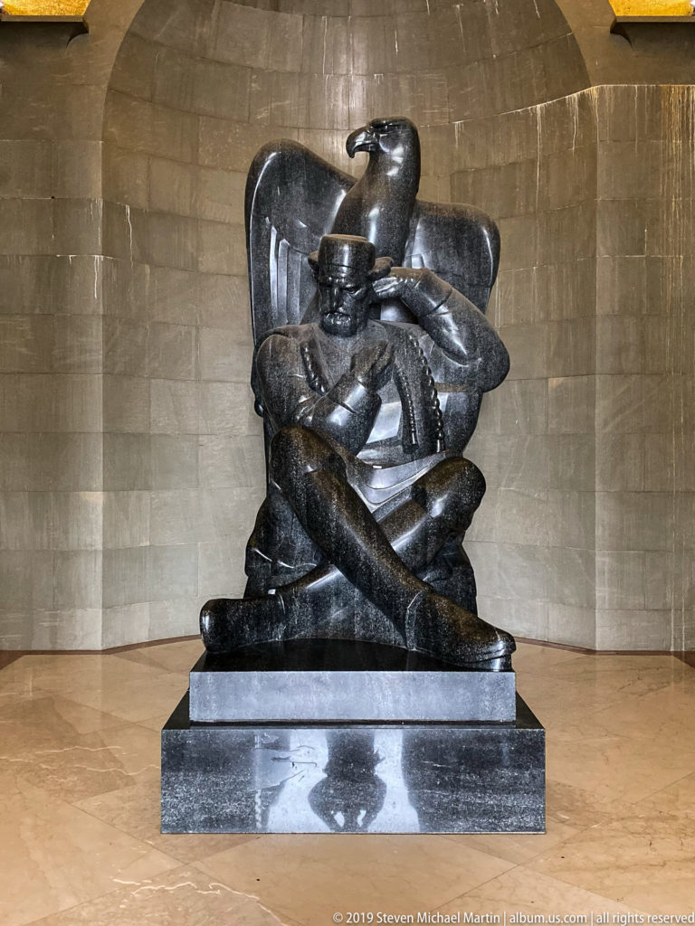 Full frontal view of the seated dark-stone Njegoš statue with crossed legs and a stylized eagle rising behind.