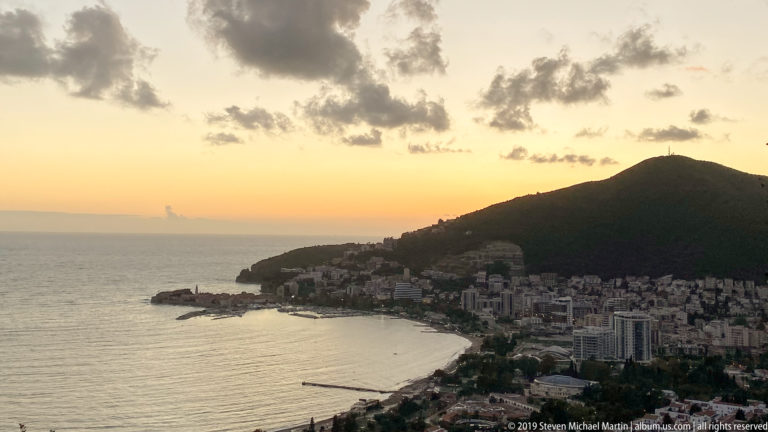 Elevated sunset view of a Montenegrin coastal town around a bay, with the Adriatic Sea and dark mountain ridge beyond.