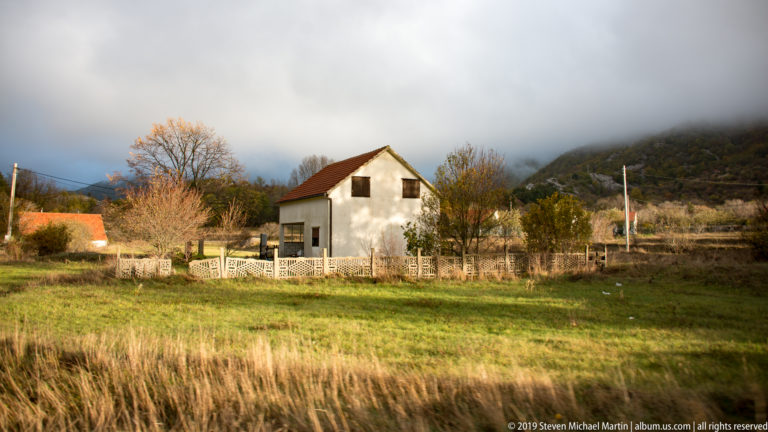 White two-story house with red roof behind a low fence, surrounded by pasture and autumn trees under misty hills.