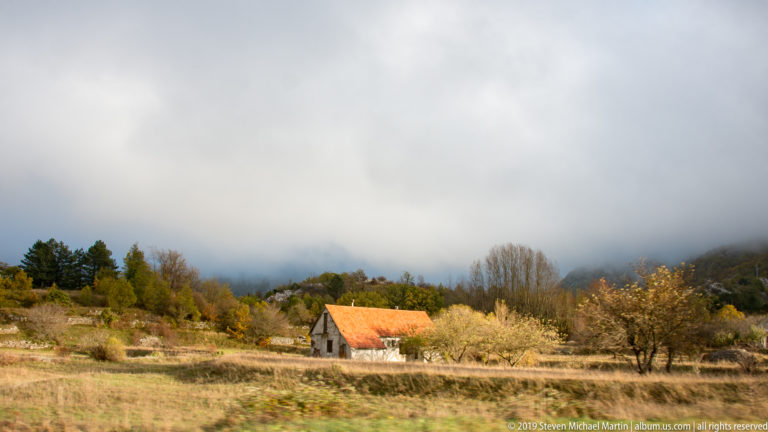 Small red-roofed rural house in pale fields and orchard trees beneath low cloud over nearby hills.