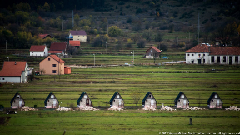 Row of small gabled stone structures across green fields with red-roofed village houses behind them.