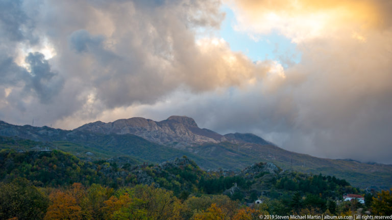 Layered Lovćen ridges and autumn woodland beneath dramatic gray and gold clouds with a patch of blue sky.